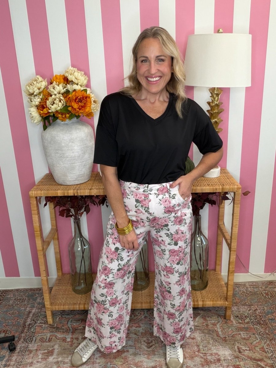 A woman stands in a pink and white striped room wearing a black short-sleeve V-neck top and white wide-leg pants with a pink floral print. She is standing in front of a wooden console table with a vase of flowers and a lamp.