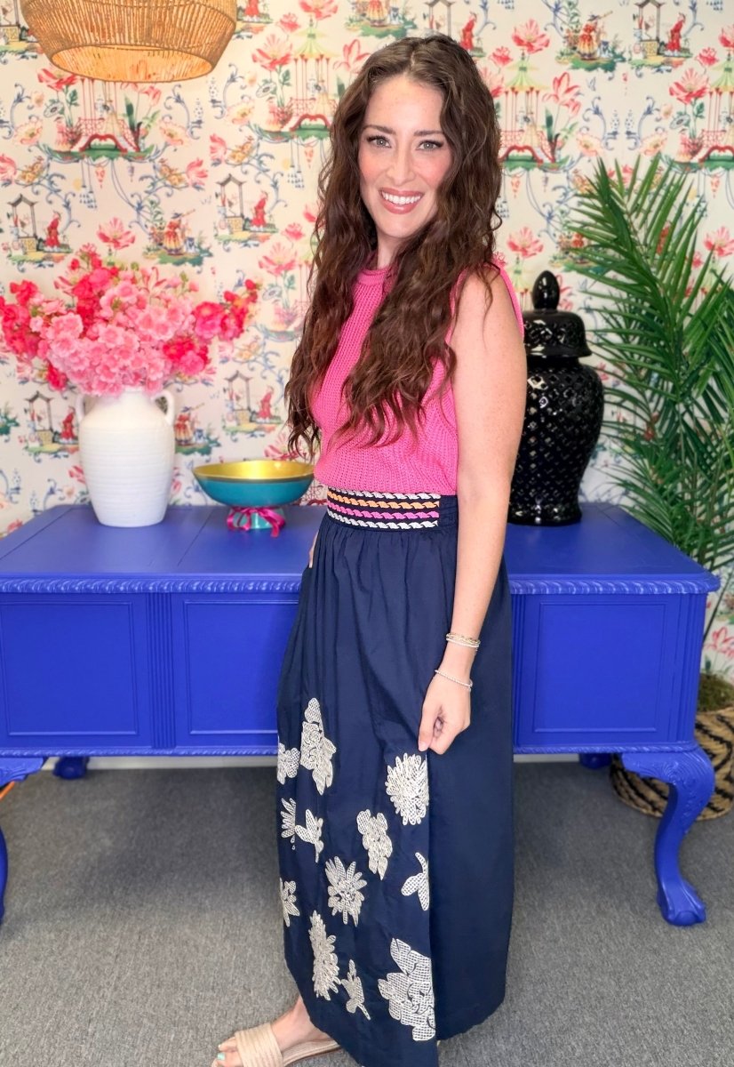 A woman wearing a sleeveless pink top and a navy maxi skirt with white floral embroidery, standing in front of a blue cabinet and floral wallpaper.