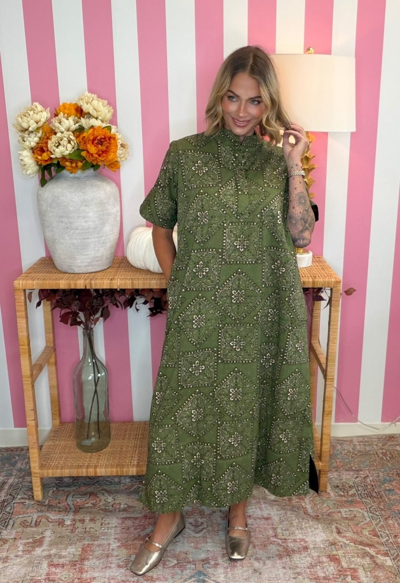 A woman models a short-sleeved olive green midi dress with intricate silver beaded geometric patterns, standing in front of a pink and white striped wall next to a wooden console table with a vase of flowers.