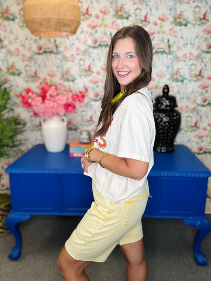 Woman in a white shirt and yellow shorts standing in a room with floral wallpaper and a blue table.