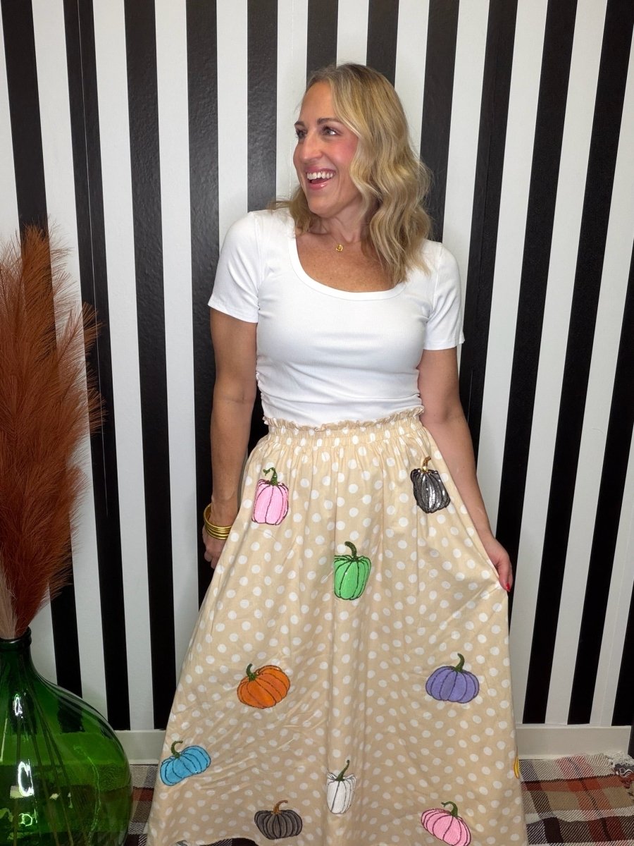 A woman wearing a white short-sleeve scoop-neck t-shirt and a beige polka-dot midi skirt decorated with colorful embroidered pumpkins, standing in front of a black and white striped wall.
