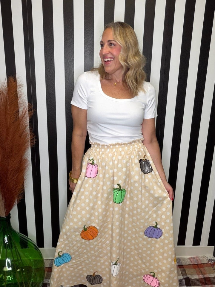 A woman wearing a white short-sleeve scoop-neck t-shirt and a beige polka-dot midi skirt decorated with colorful embroidered pumpkins, standing in front of a black and white striped wall.
