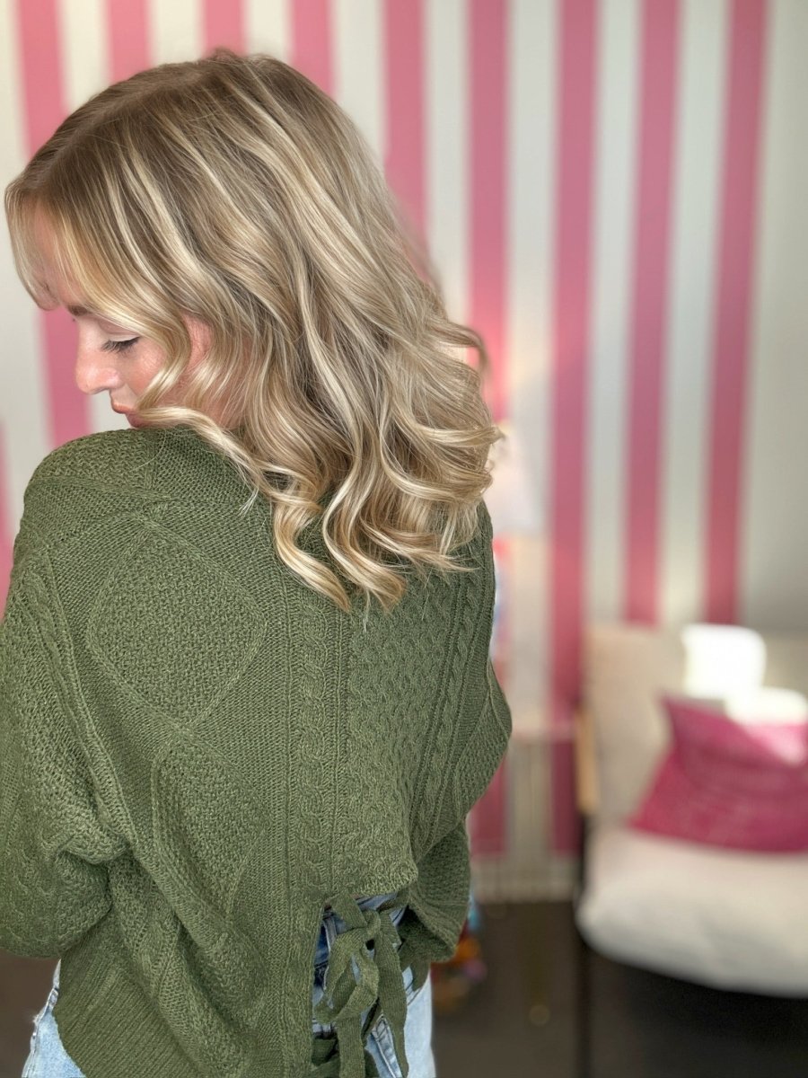 A woman with blonde wavy hair is shown from the back wearing an olive green cable-knit sweater with a tie detail at the back, set against a pink and white striped wall.