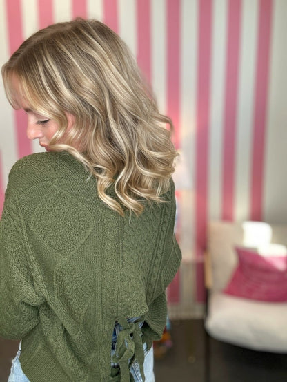 A woman with blonde wavy hair is shown from the back wearing an olive green cable-knit sweater with a tie detail at the back, set against a pink and white striped wall.