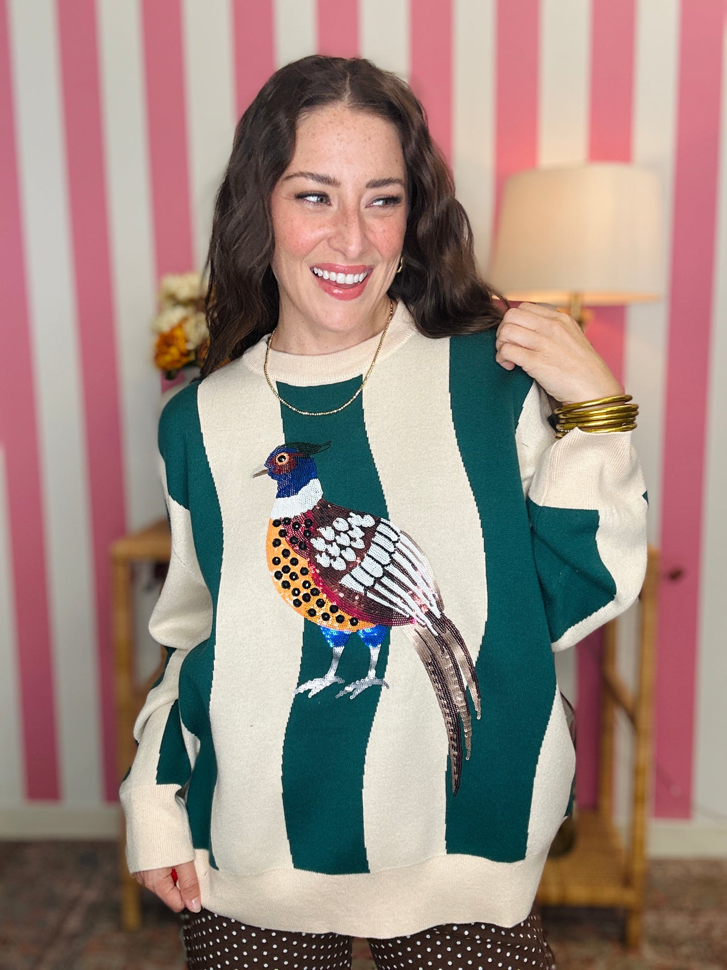A woman wearing a green and cream striped sweater with a colorful pheasant bird graphic on the front, standing in front of a pink and white striped wall.