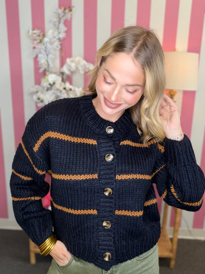 A woman wearing a navy blue knit cardigan sweater with horizontal mustard yellow stripes and gold buttons, standing in front of a pink and white striped wall.