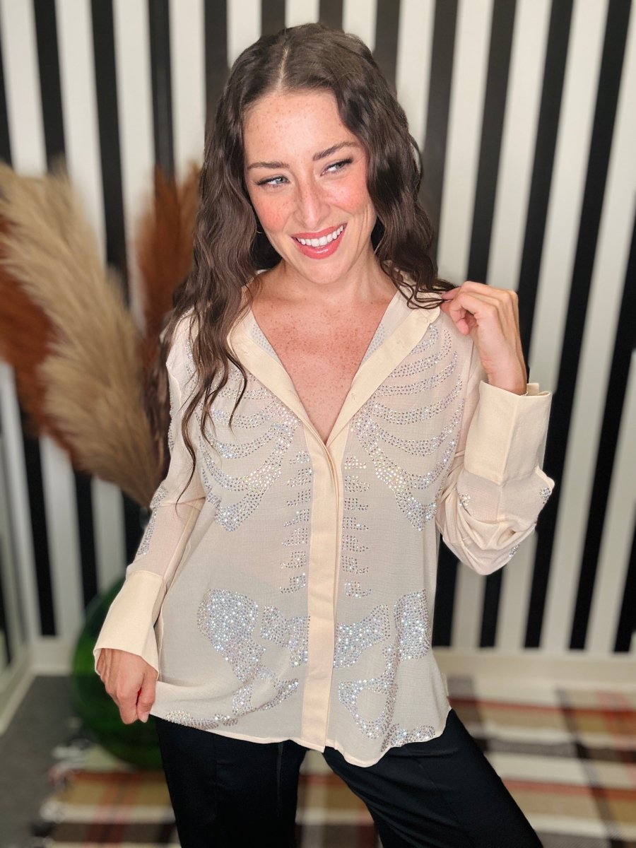 A woman wearing a beige long-sleeve blouse with silver rhinestone embellishments, paired with black pants, posing in front of a black and white striped background.