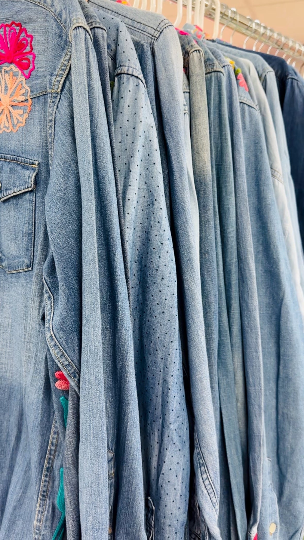 A close-up of several blue denim jackets hanging on a rack, with one jacket on the left featuring colorful floral embroidery on the back pocket.