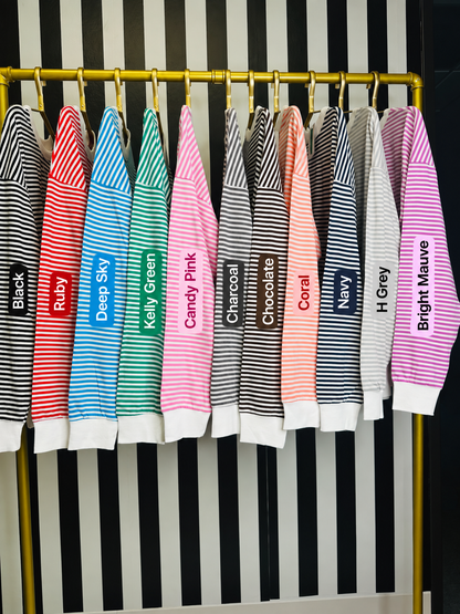 A set of colorful striped knit long-sleeve tops hanging on a gold rack against a black and white striped background. Each top has a different color and name printed on the front, including Black, Ruby, Deep Sky, Kelly Green, Candy Pink, Charcoal, Chocolate, Coral, Navy, H Grey, and Bright Mauve.