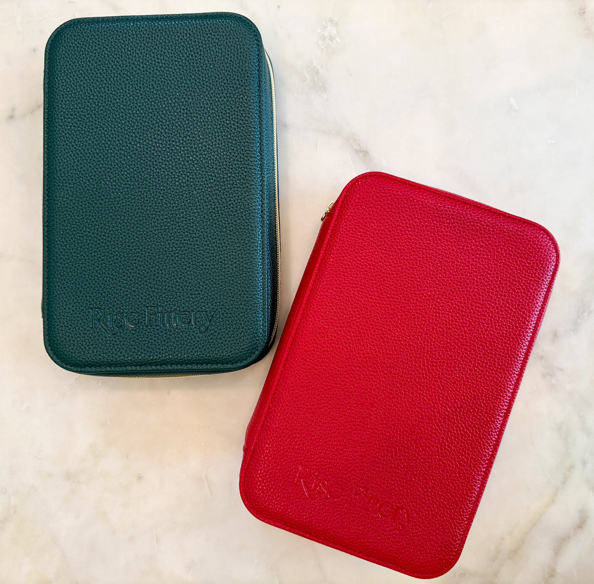 Two textured zippered carrying cases on a light marble surface: one dark teal-green and one bright red, both embossed with the brand name 'Rise Up Way'.