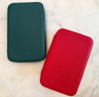 Two textured zippered carrying cases on a light marble surface: one dark teal-green and one bright red, both embossed with the brand name 'Rise Up Way'.