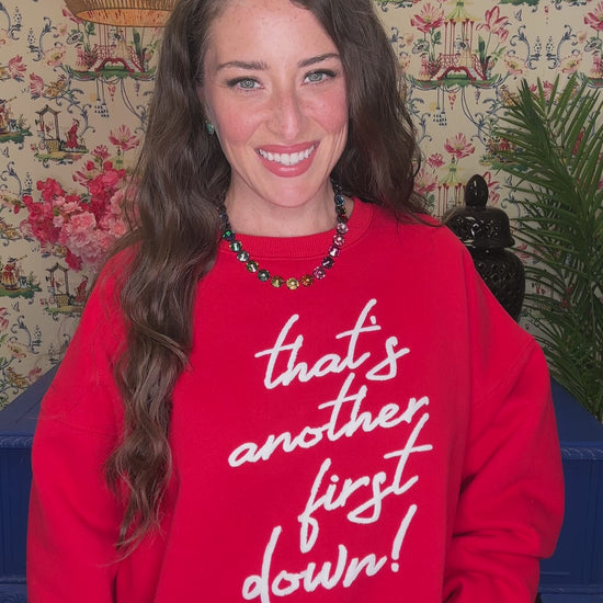 A woman smiling while wearing a red crewneck sweatshirt with the phrase 'that's another first down!' printed in white script across the front, paired with blue jeans in a decorated indoor setting.