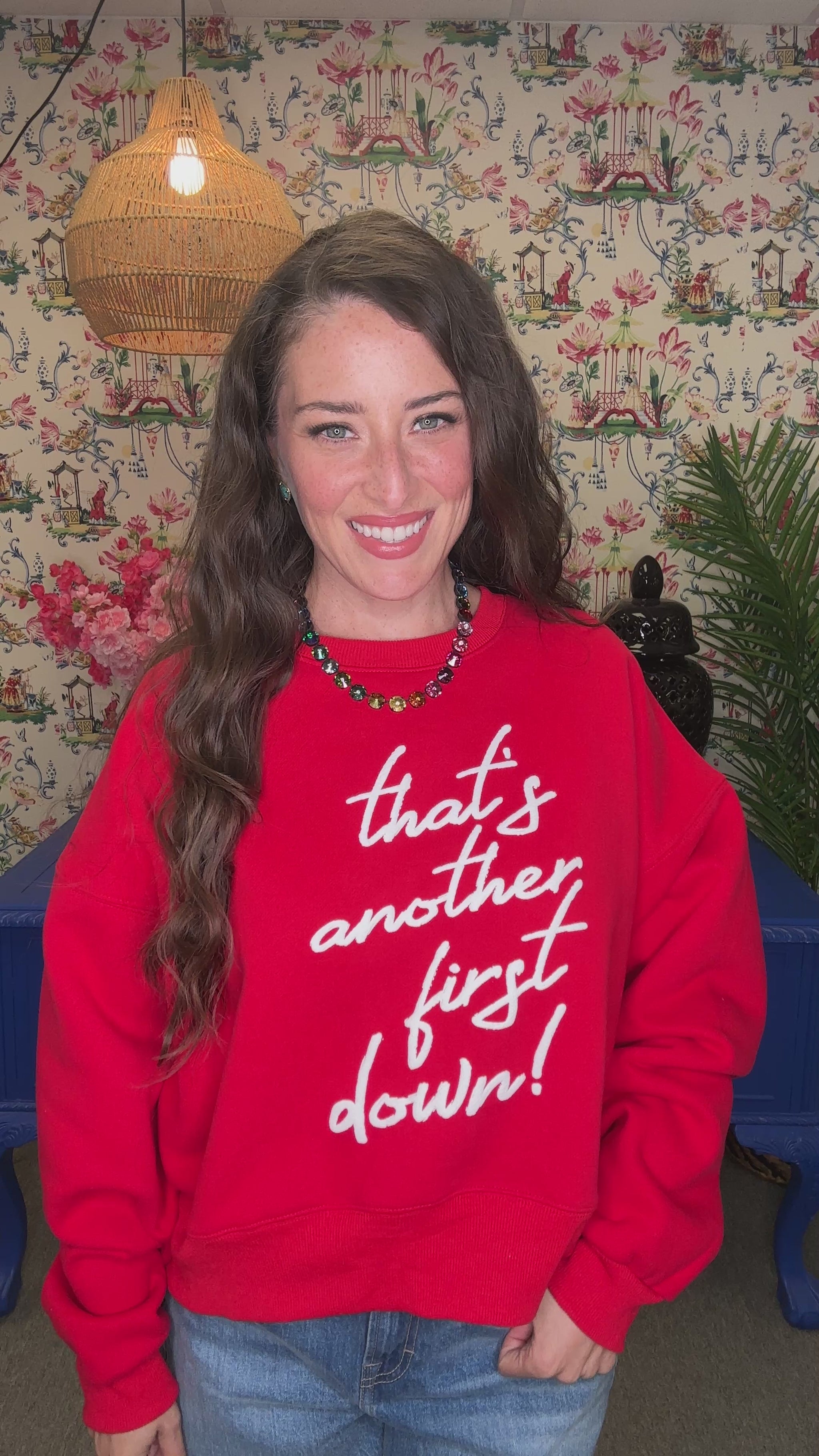 A woman smiling while wearing a red crewneck sweatshirt with the phrase 'that's another first down!' printed in white script across the front, paired with blue jeans in a decorated indoor setting.
