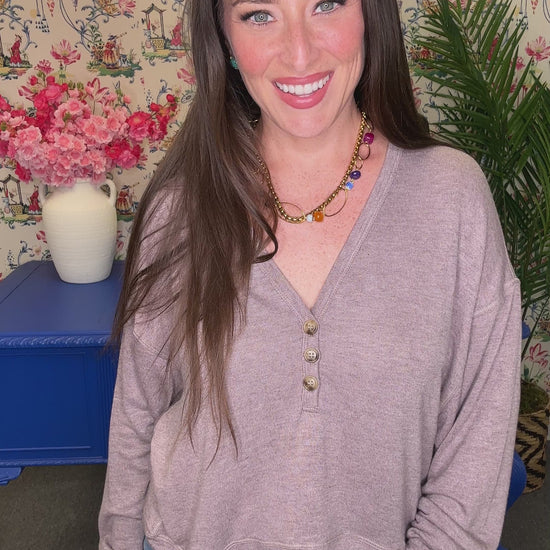 A smiling woman with long brown hair wearing a light mauve-purple long-sleeve knit top and blue jeans, standing in a decorated room with floral wallpaper, a blue table, and plants.