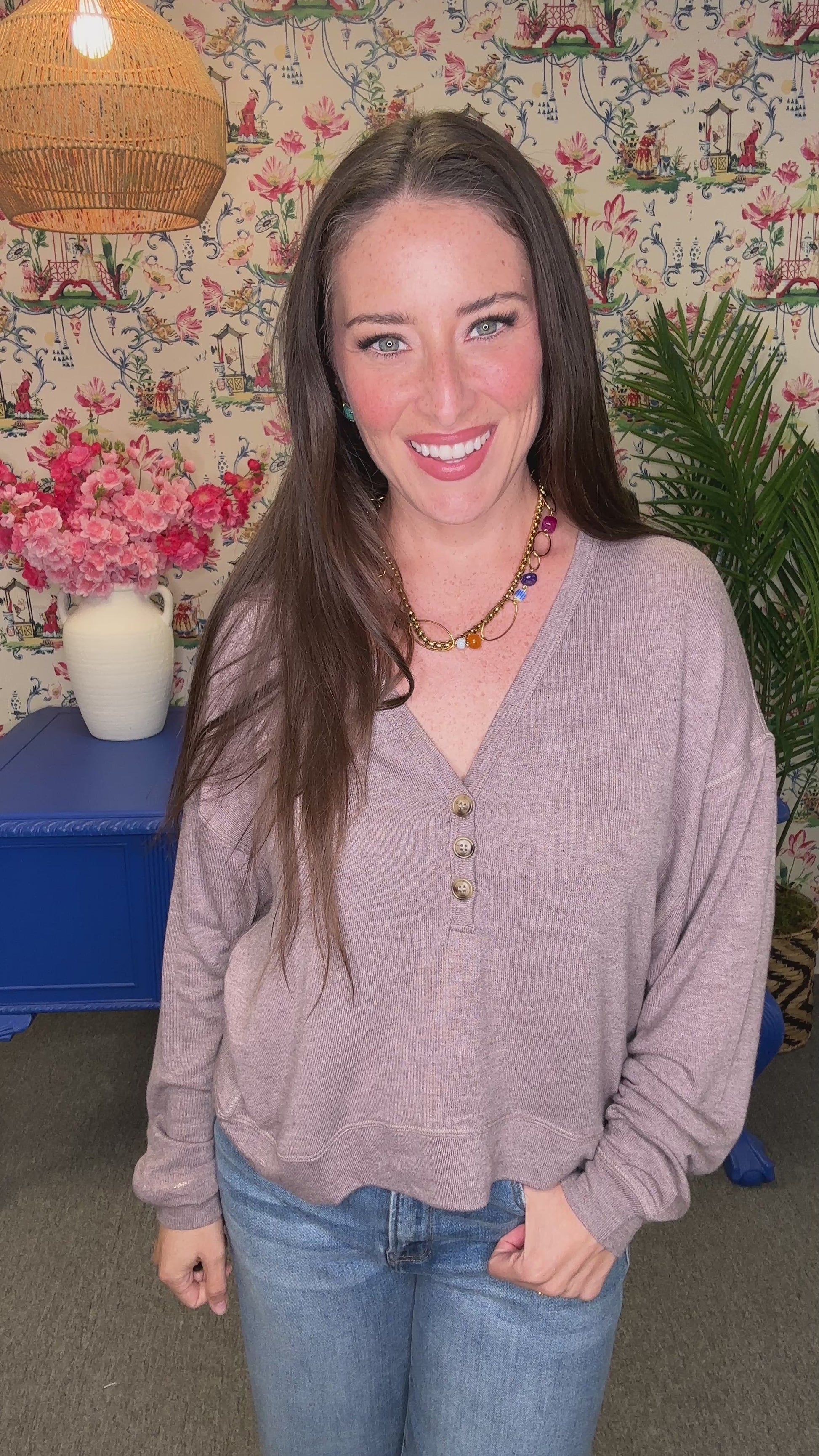 A smiling woman with long brown hair wearing a light mauve-purple long-sleeve knit top and blue jeans, standing in a decorated room with floral wallpaper, a blue table, and plants.