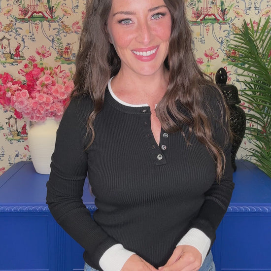 A woman with dark hair smiles while holding up a black knit polo sweater on a hanger with a white collar, wearing a taupe ribbed henley sweater and blue jeans in front of a floral wallpaper background.