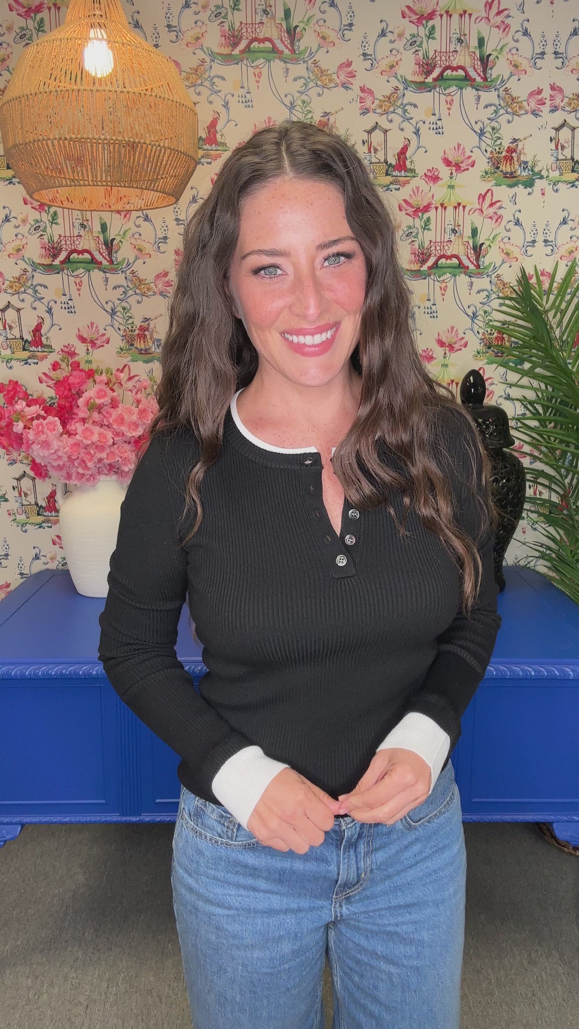 A woman with dark hair smiles while holding up a black knit polo sweater on a hanger with a white collar, wearing a taupe ribbed henley sweater and blue jeans in front of a floral wallpaper background.
