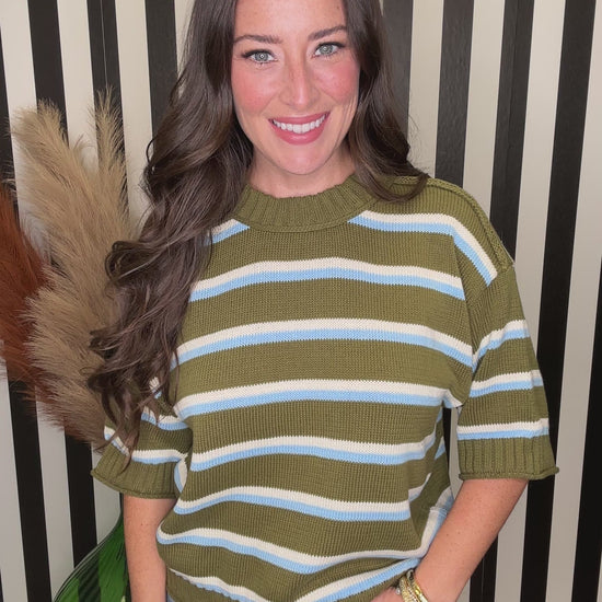 A woman with long wavy brown hair wearing a short-sleeve knit sweater with horizontal olive green, white, and light blue stripes, paired with blue jeans. She is smiling and looking down, with her hand near her hair, set against a black and white striped background.