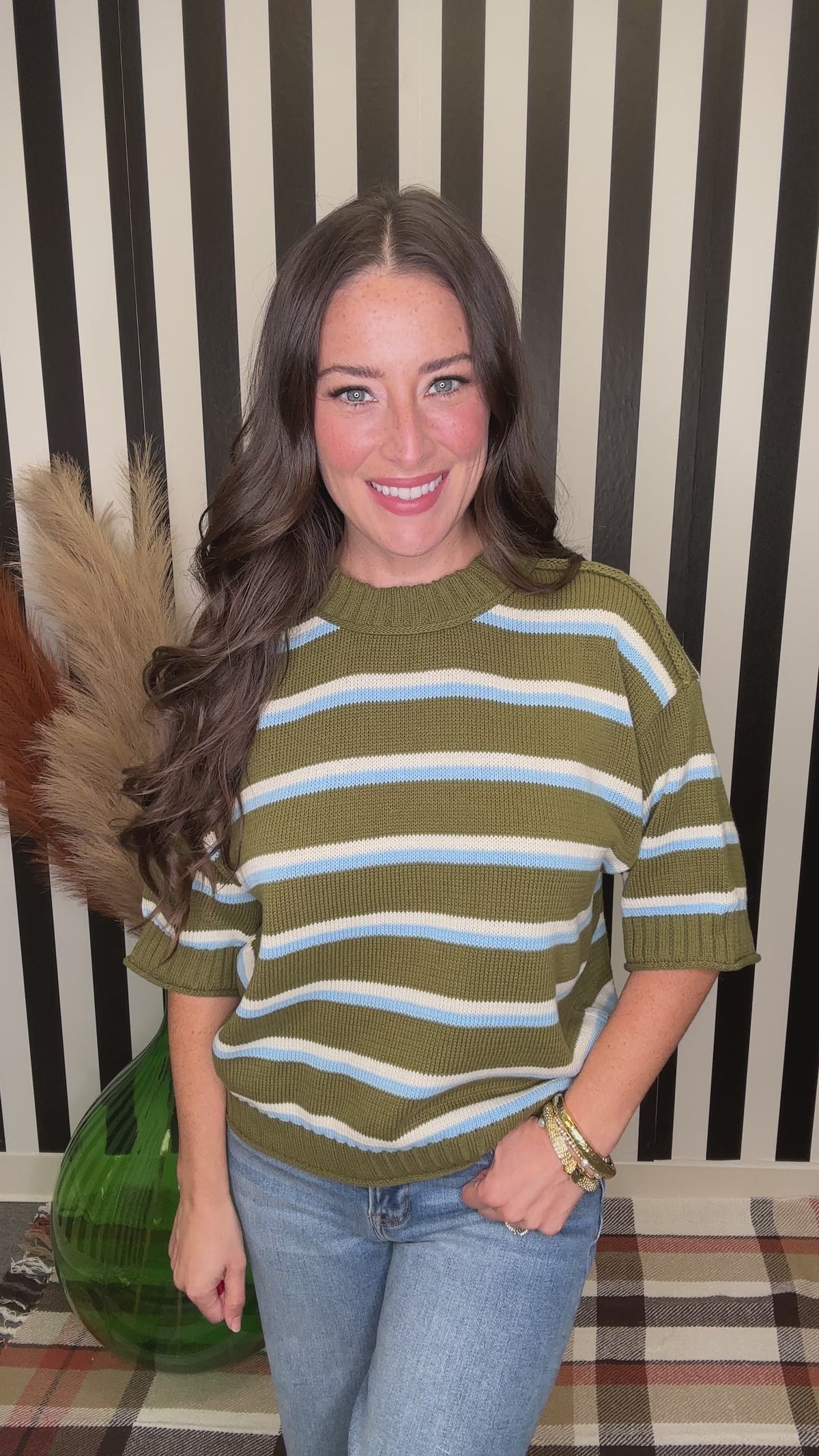 A woman with long wavy brown hair wearing a short-sleeve knit sweater with horizontal olive green, white, and light blue stripes, paired with blue jeans. She is smiling and looking down, with her hand near her hair, set against a black and white striped background.