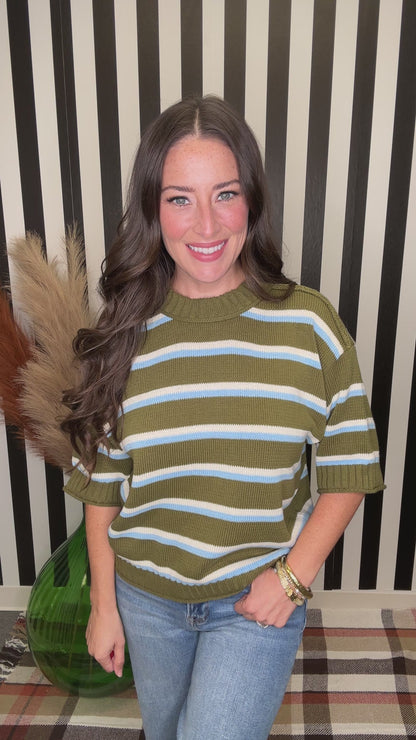 A woman with long wavy brown hair wearing a short-sleeve knit sweater with horizontal olive green, white, and light blue stripes, paired with blue jeans. She is smiling and looking down, with her hand near her hair, set against a black and white striped background.