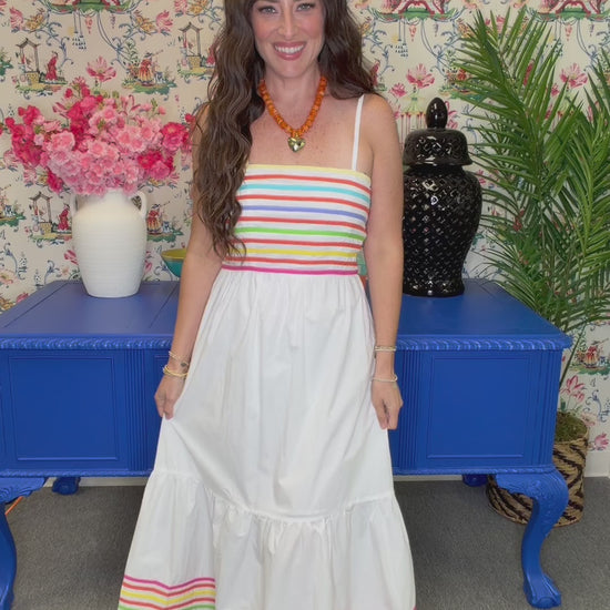 A woman modeling a white tiered maxi dress with colorful horizontal stripes at the bust and hem, standing in front of a floral wallpaper background and a blue cabinet.