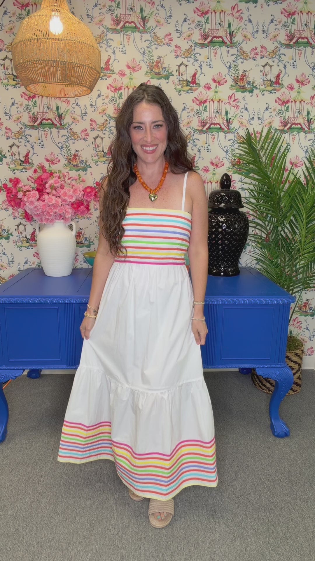 A woman modeling a white tiered maxi dress with colorful horizontal stripes at the bust and hem, standing in front of a floral wallpaper background and a blue cabinet.