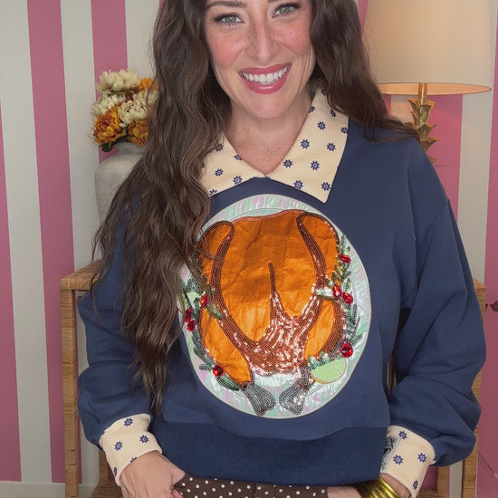A woman wearing a navy blue sweatshirt with a large roasted turkey graphic on the front, a cream polka-dot collar, and cream polka-dot cuffs, paired with brown polka-dot pants, posed in front of a pink and white striped wall.