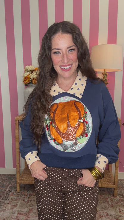 A woman wearing a navy blue sweatshirt with a large roasted turkey graphic on the front, a cream polka-dot collar, and cream polka-dot cuffs, paired with brown polka-dot pants, posed in front of a pink and white striped wall.