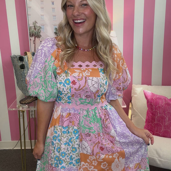 A woman modeling a colorful patchwork maxi dress with puff sleeves, standing in front of a pink and white striped wall. She is wearing a gold necklace and sandals, and the background includes a side table with a denim tote bag, a framed print, and a white chair with a pink pillow.