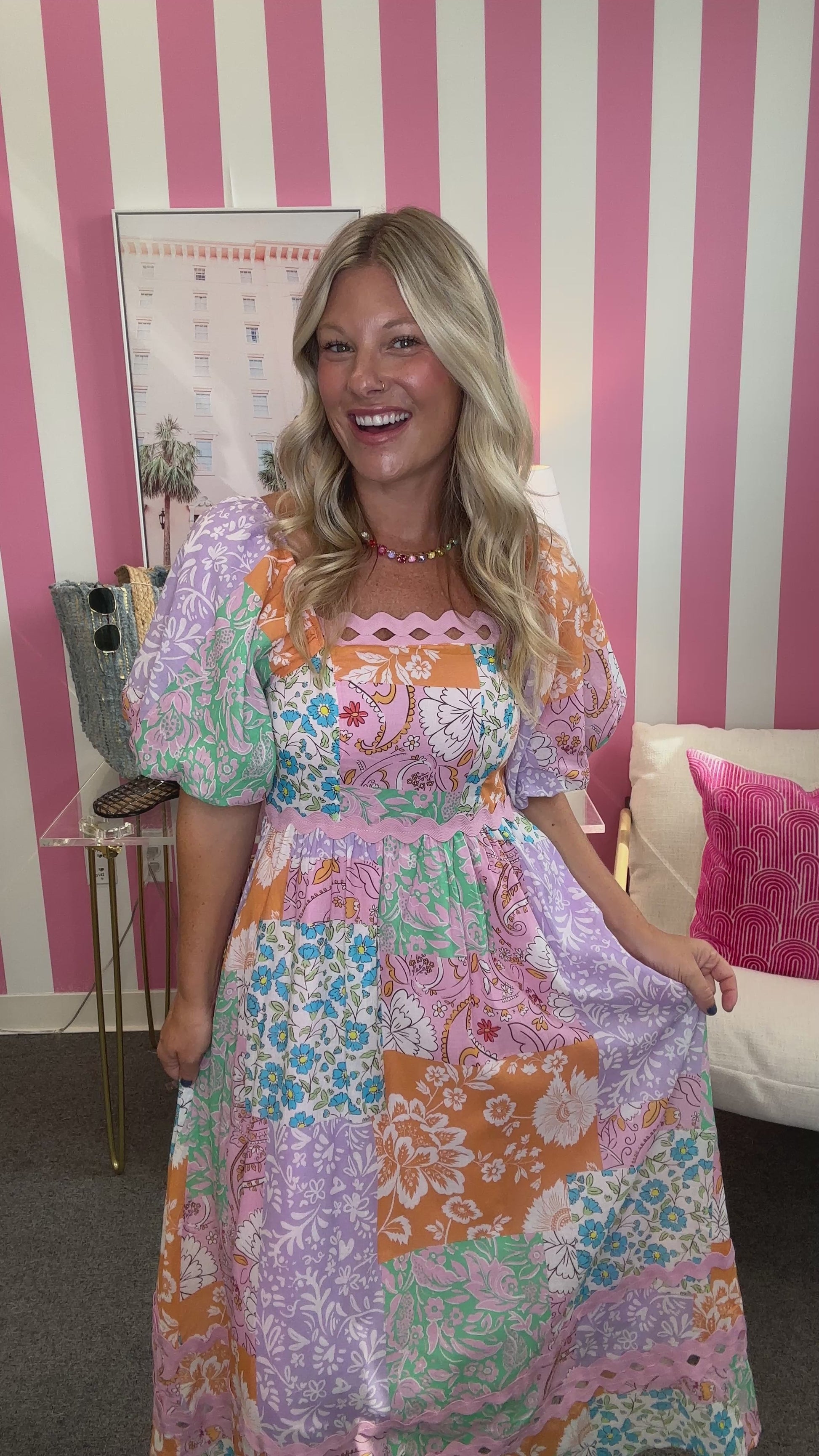 A woman modeling a colorful patchwork maxi dress with puff sleeves, standing in front of a pink and white striped wall. She is wearing a gold necklace and sandals, and the background includes a side table with a denim tote bag, a framed print, and a white chair with a pink pillow.