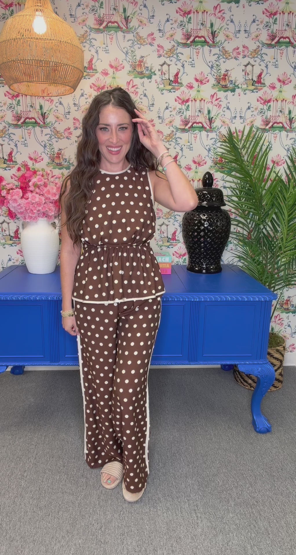 A woman modeling a brown and white polka dot sleeveless maxi dress with white piping, standing in front of a blue cabinet and floral wallpaper.
