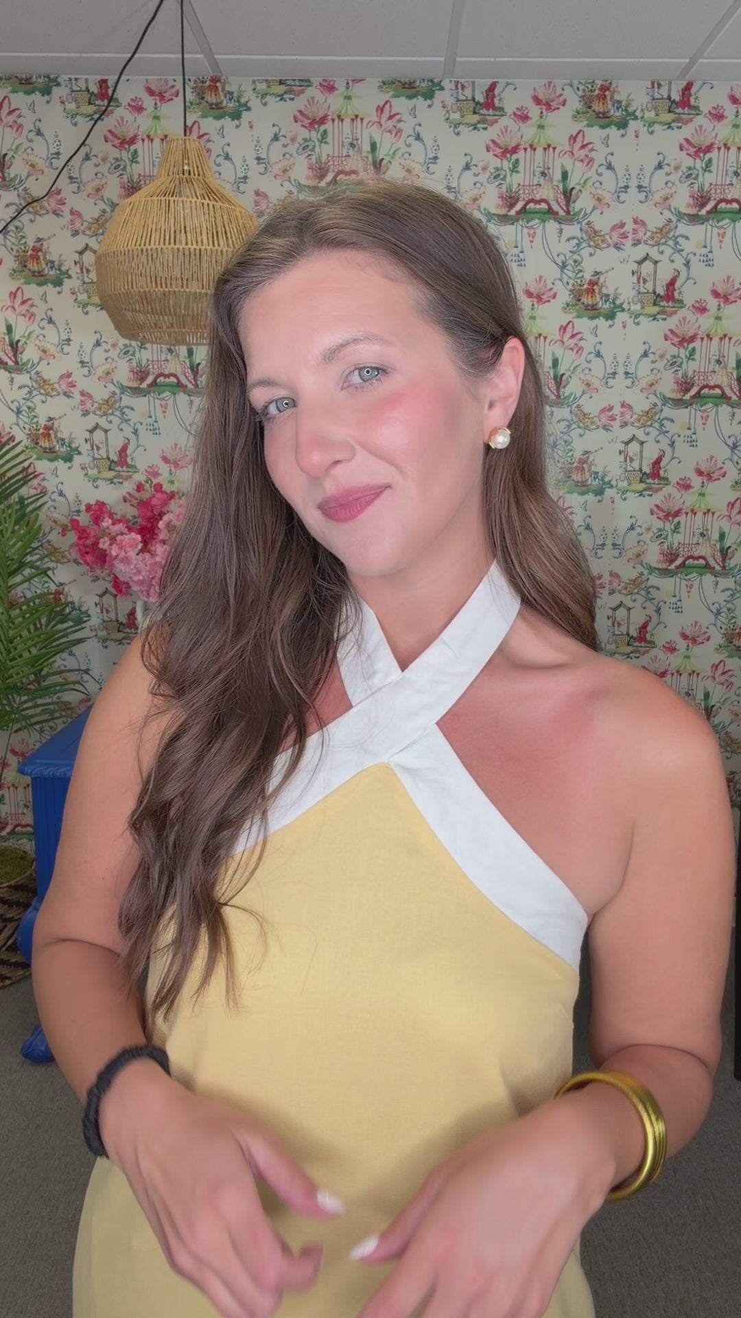 Woman in a yellow dress standing in front of a colorful floral-patterned wall with decorative vases and flowers.