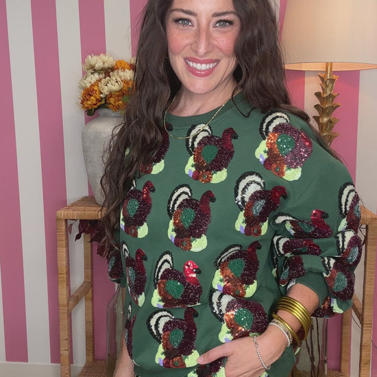 A woman modeling a green sequined long-sleeve top with a colorful turkey pattern, standing in front of a pink and white striped wall with a wooden side table and lamp in the background.