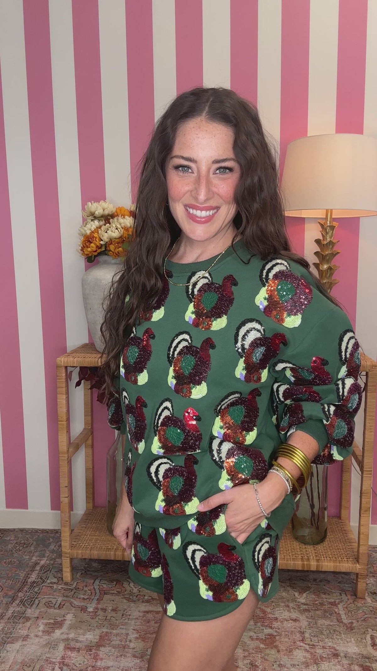 A woman modeling a green sequined long-sleeve top with a colorful turkey pattern, standing in front of a pink and white striped wall with a wooden side table and lamp in the background.