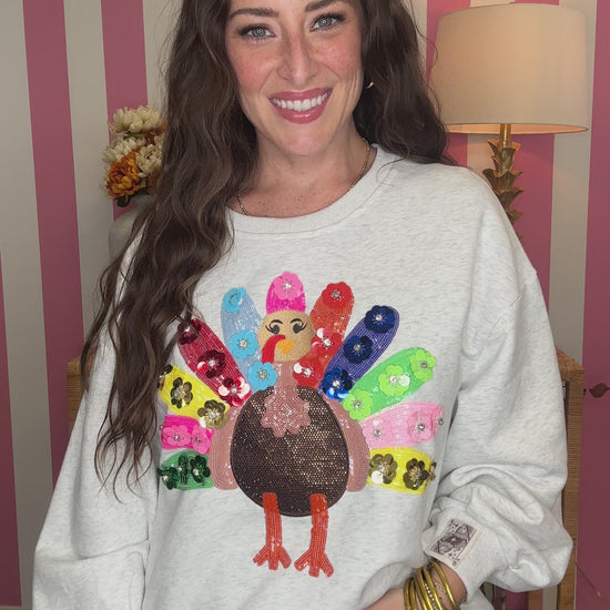 A woman smiling while wearing a light gray crewneck sweatshirt with a colorful sequined turkey graphic on the front, paired with brown polka dot pants, set against a pink and white striped wall background.