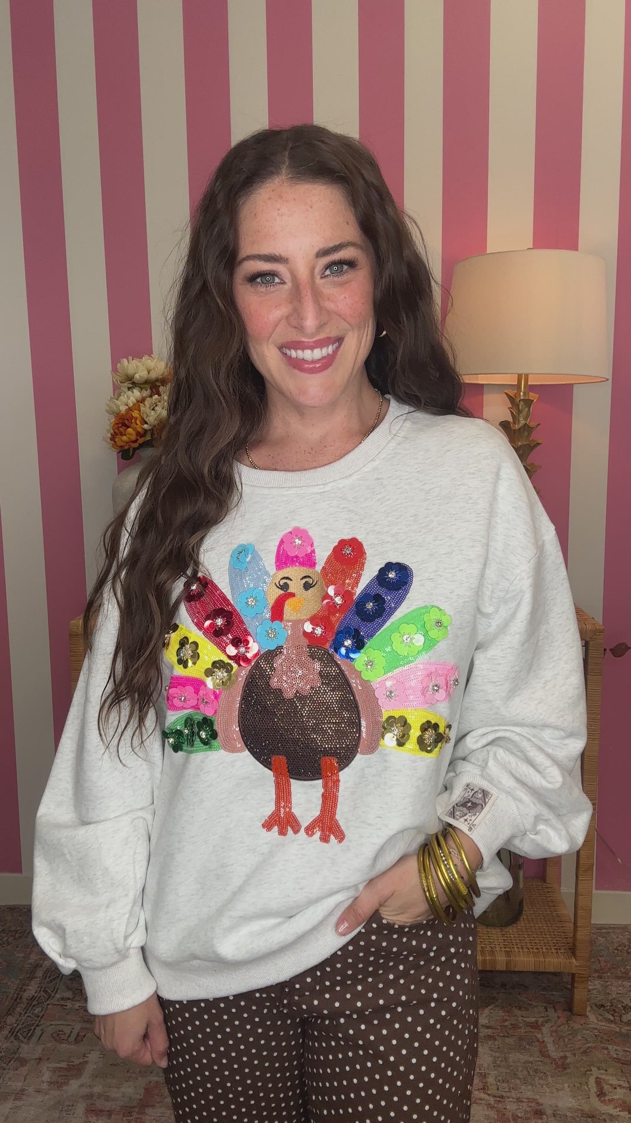 A woman smiling while wearing a light gray crewneck sweatshirt with a colorful sequined turkey graphic on the front, paired with brown polka dot pants, set against a pink and white striped wall background.