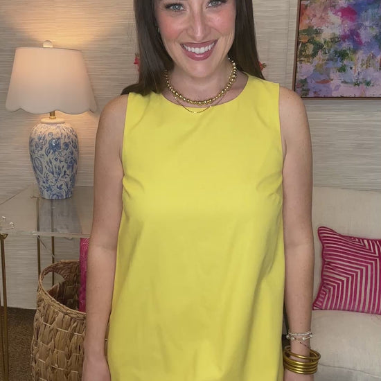 Woman in a yellow dress standing in a living room with a lamp and colorful painting in the background.
