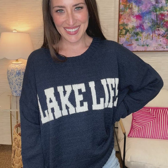 Woman holding a blue 'Lake Life' sweater in a room with colorful decor.
