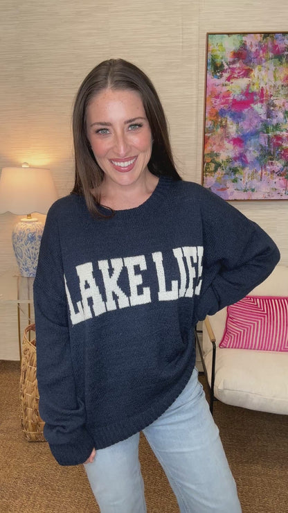 Woman holding a blue 'Lake Life' sweater in a room with colorful decor.