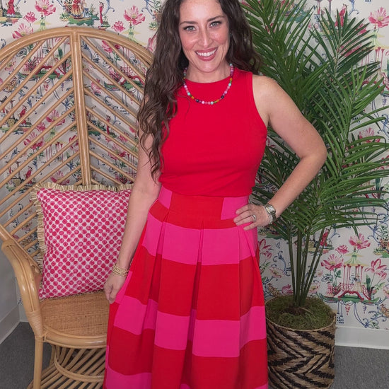 Woman in a red and pink dress standing next to a wicker chair with a pink cushion and a plant against a patterned wall.