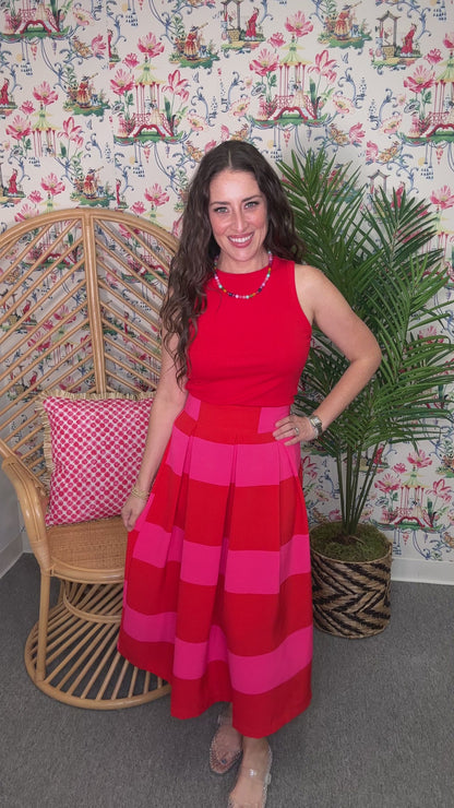 Woman in a red and pink dress standing next to a wicker chair with a pink cushion and a plant against a patterned wall.