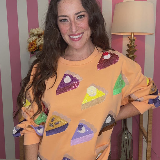 A woman smiling while wearing a peach-colored sweatshirt covered in sequined pie-slice patches, posed in front of a pink and white striped wall.