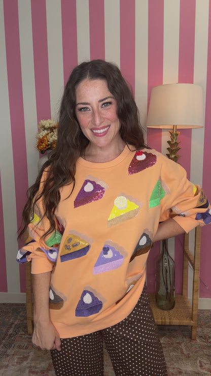 A woman smiling while wearing a peach-colored sweatshirt covered in sequined pie-slice patches, posed in front of a pink and white striped wall.