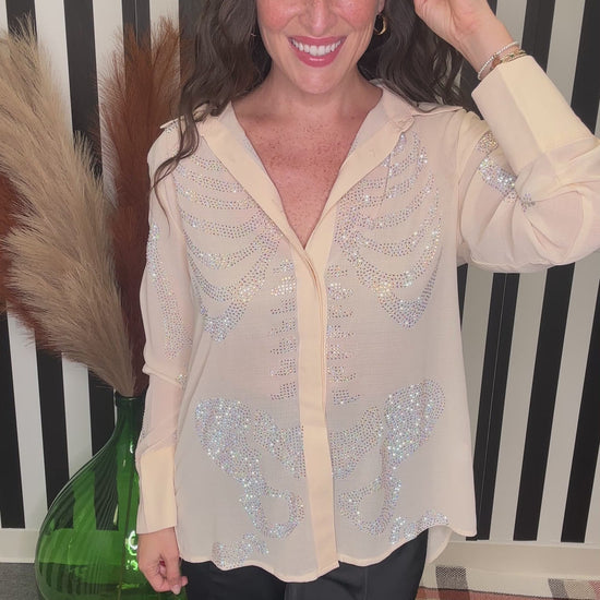 A woman wearing a sheer beige button-up blouse with silver sequin embellishments, paired with black pants, standing in front of a black and white striped background.