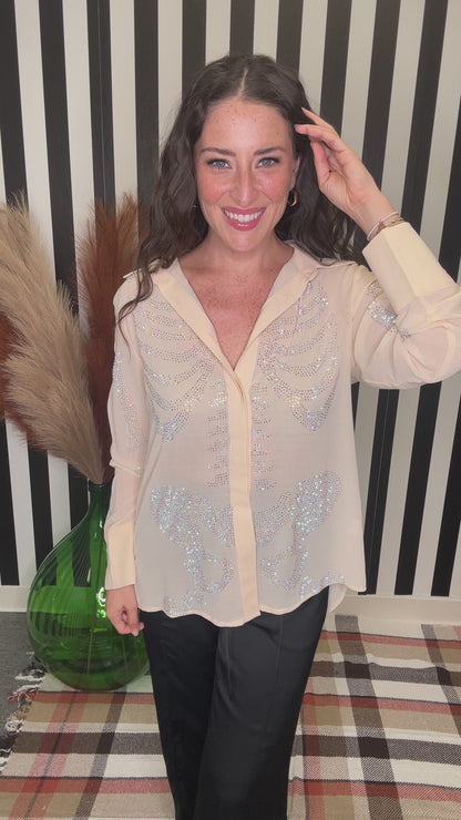 A woman wearing a sheer beige button-up blouse with silver sequin embellishments, paired with black pants, standing in front of a black and white striped background.