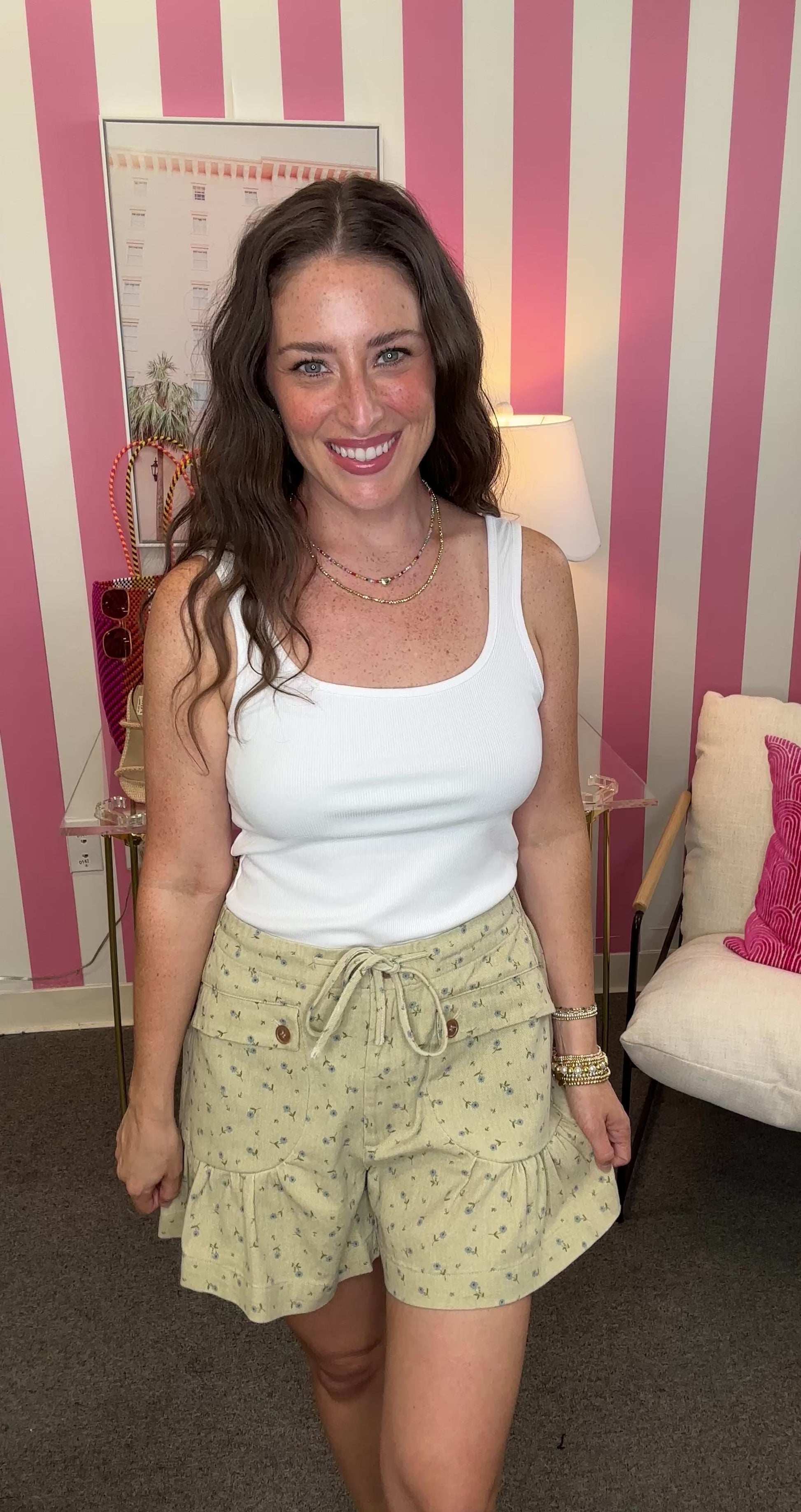 Woman wearing a white tank top and light green shorts in a room with pink walls.