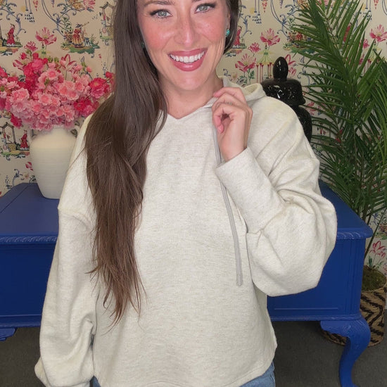 A woman with long brown hair smiling and looking back over her shoulder, wearing a light heather gray hoodie and blue jeans, standing in front of a floral wallpaper background with a blue table.