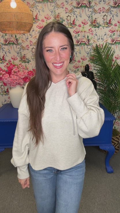 A woman with long brown hair smiling and looking back over her shoulder, wearing a light heather gray hoodie and blue jeans, standing in front of a floral wallpaper background with a blue table.