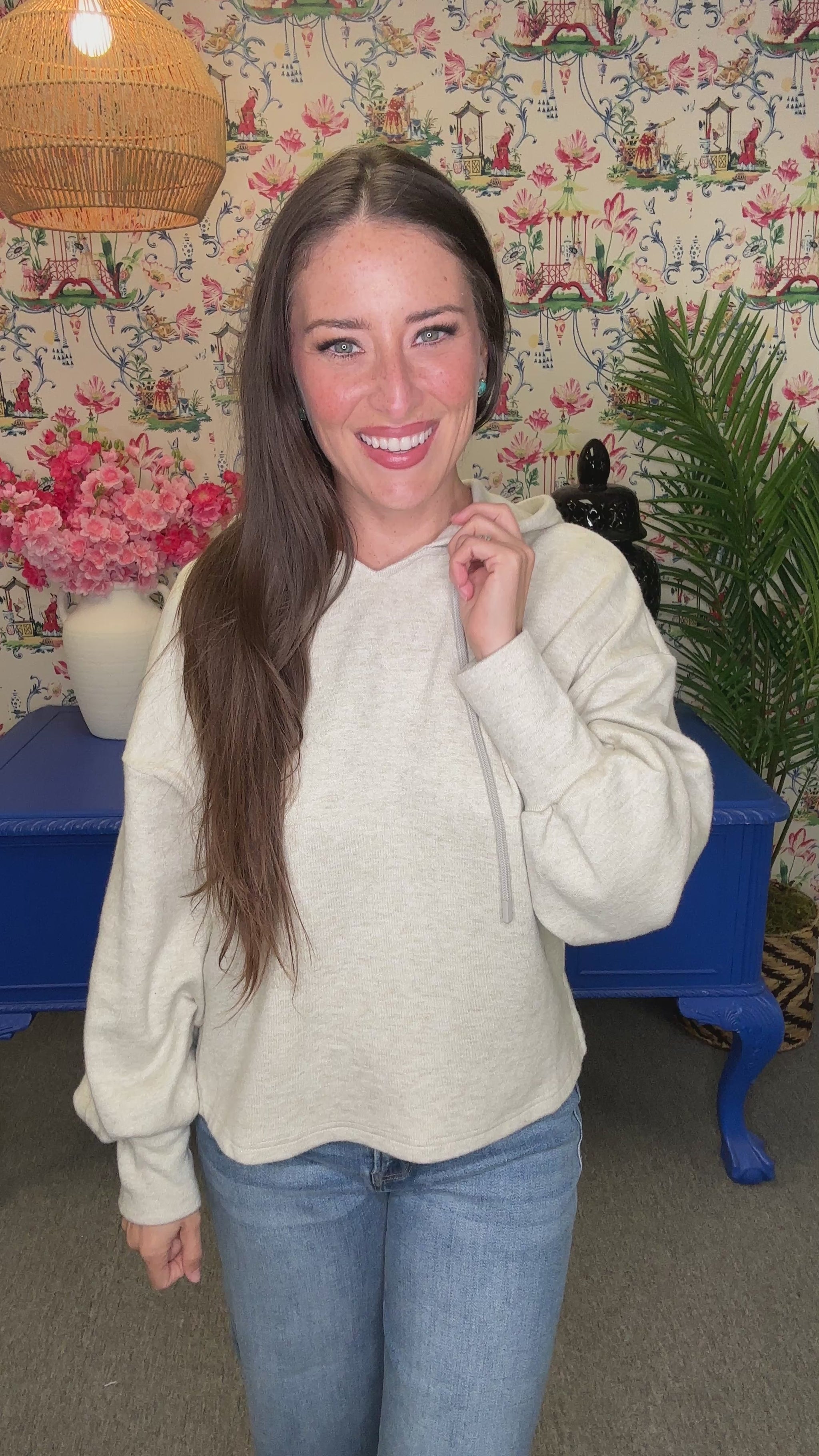 A woman with long brown hair smiling and looking back over her shoulder, wearing a light heather gray hoodie and blue jeans, standing in front of a floral wallpaper background with a blue table.