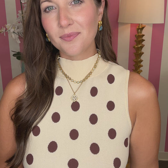 A woman wearing a sleeveless cream top with brown polka dots and white pants, standing in a room with pink and white striped walls, a lamp, and a side table.

